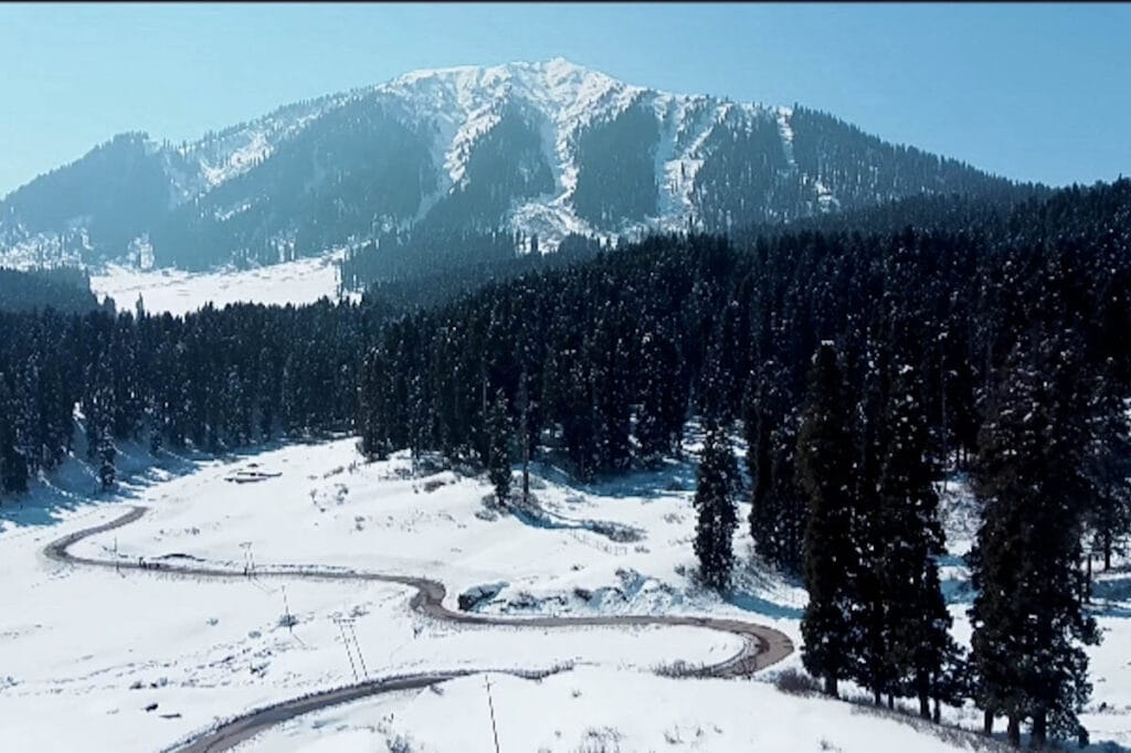 Arial view of curved road in snow covered meadow of Doodhpathri surrounded by pine forests and snow-cladded mountain in background
